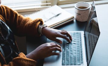 Students use online classes to stay ahead at SLCC Black woman typing on a black and silver laptop with notebook and coffee mug resting on a table