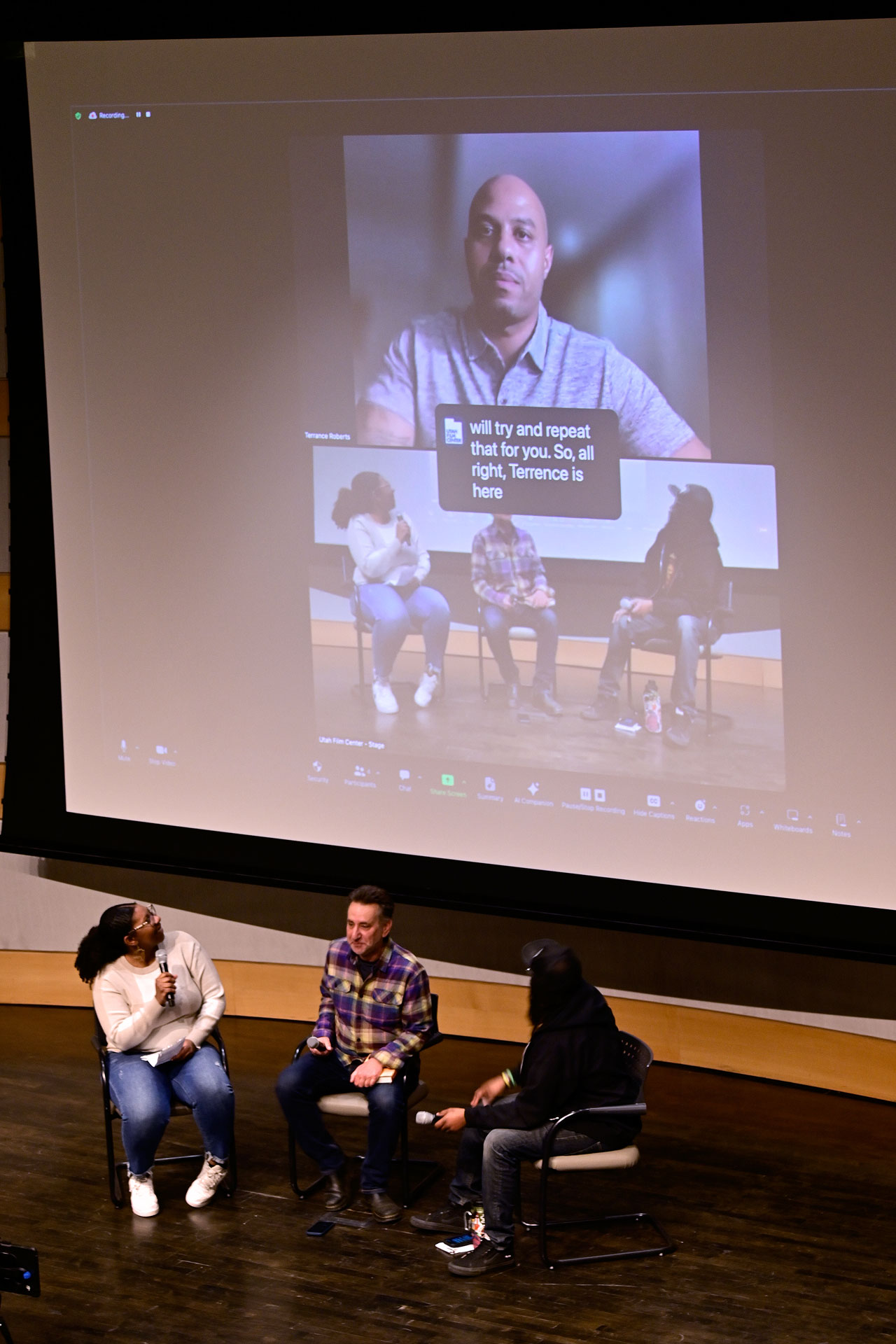 Three panelists seated on stage underneath large projection screen showing Terrance Roberts