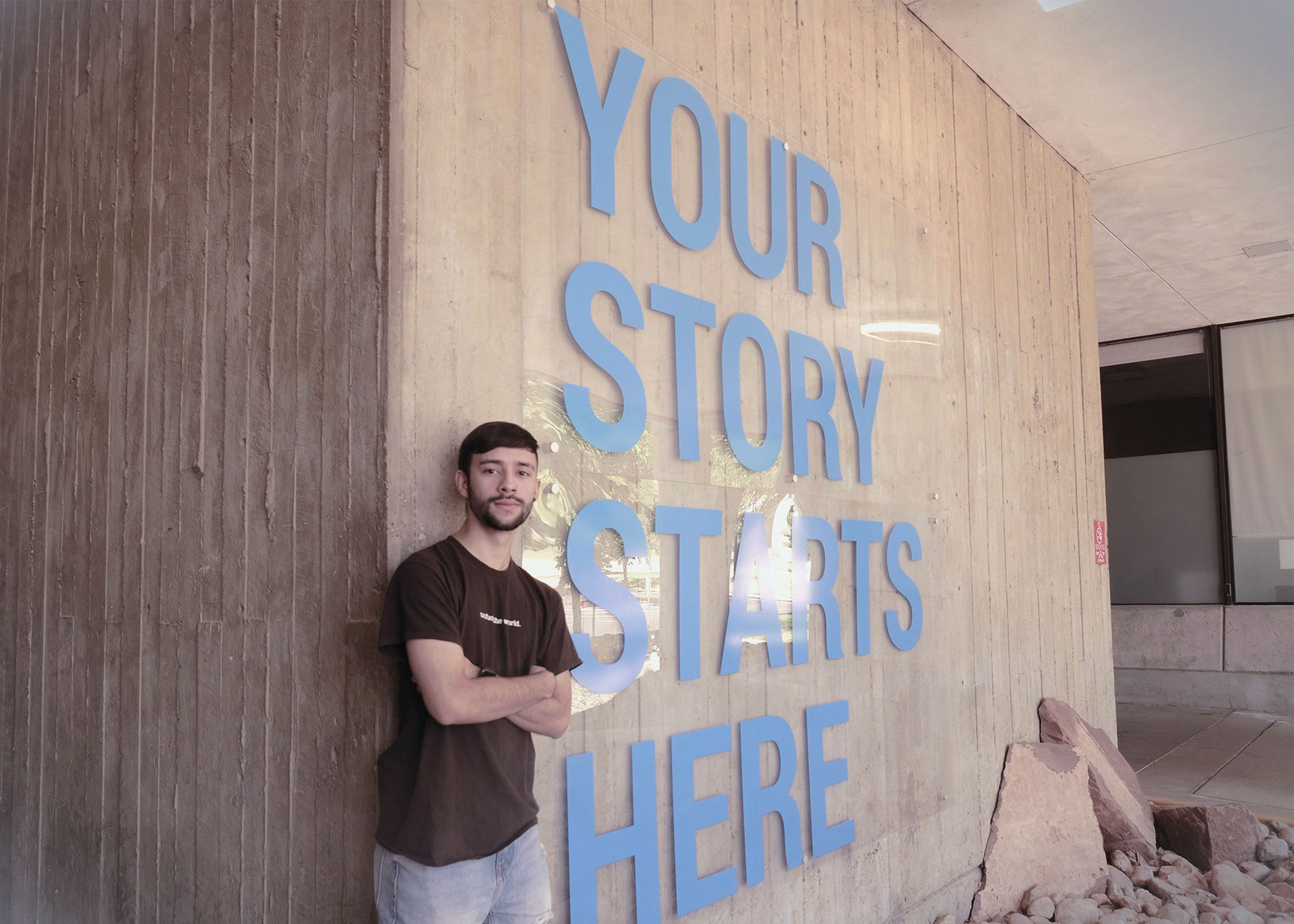 Emmanuel standing by a cement wall with text that reads, "Your story starts here"