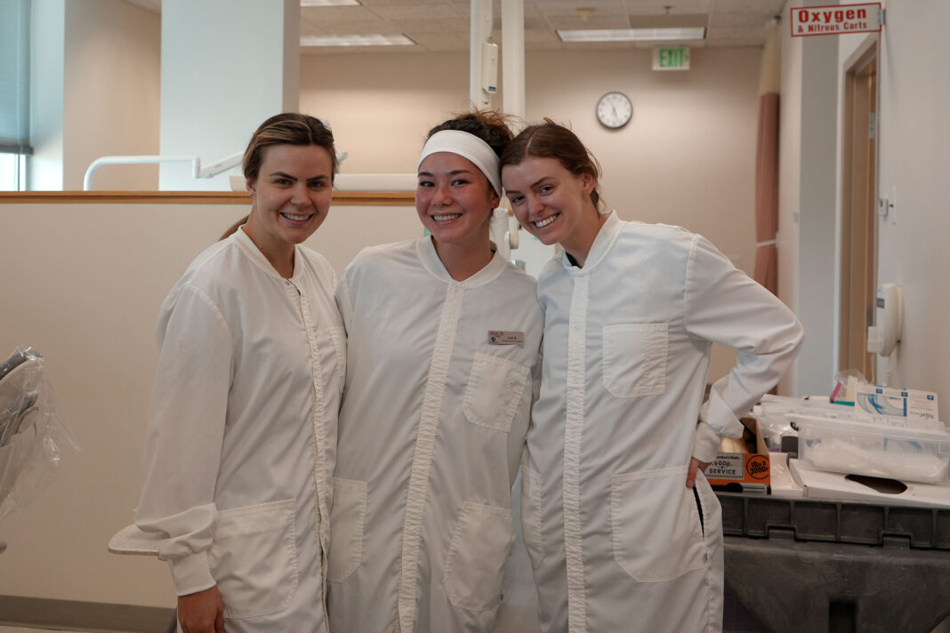 Three dental hygiene students wearing white jumpsuits in the clinic