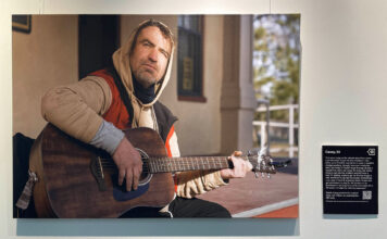 Stories through photographs, and the humanity therein Photo of Casey playing a guitar with a description of his story being displayed on a wall