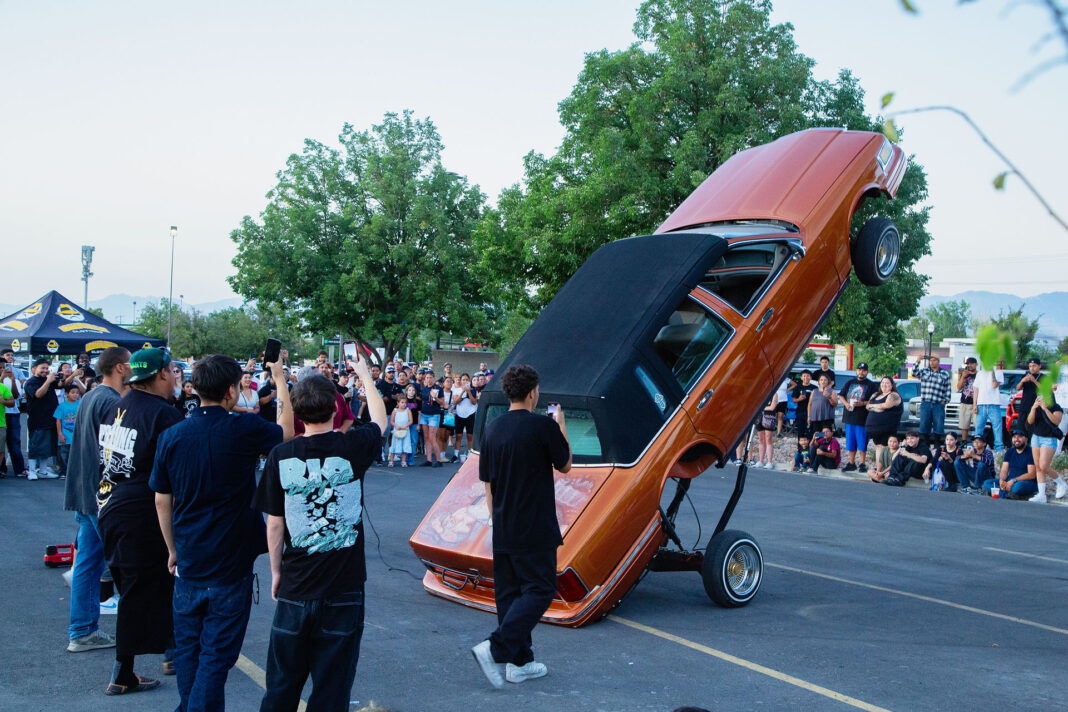 Spectators surrounding an orange lowrider hopping on its rear wheels and bumper