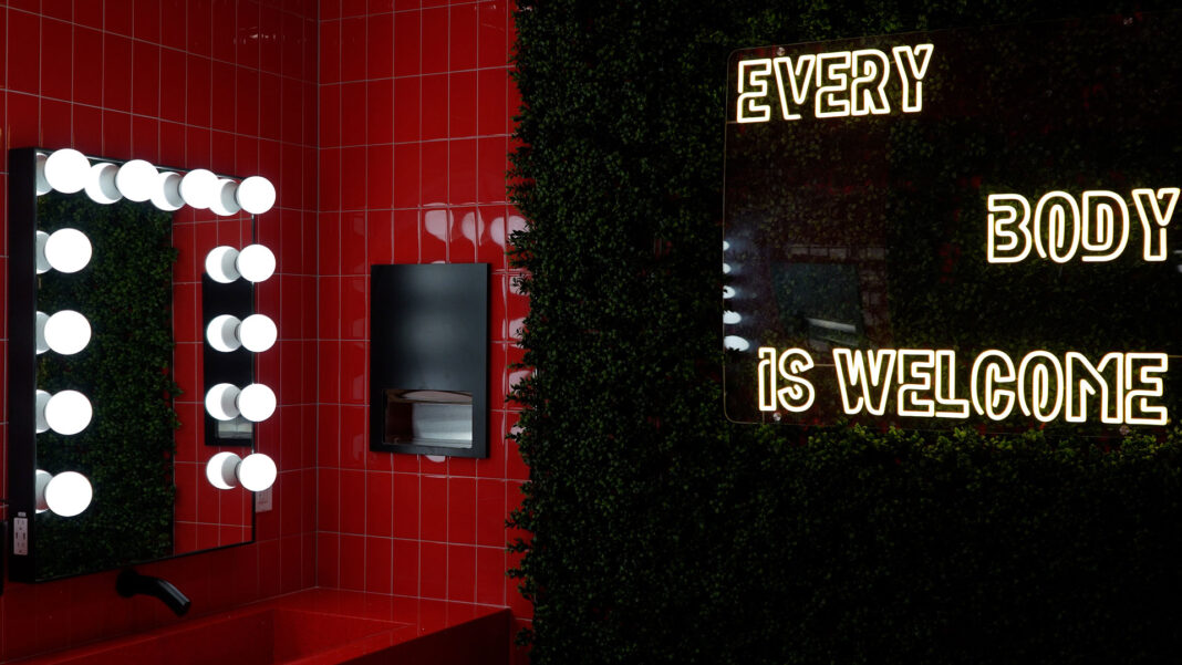 Light bulbs illuminate a mirror on a red tile wall next to a halogen sign that reads "every body is welcome"