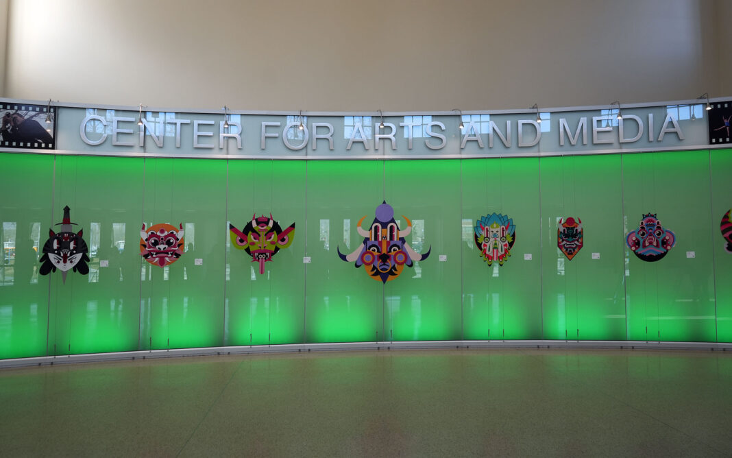 Row of masks made of mixed media lining a LED wall