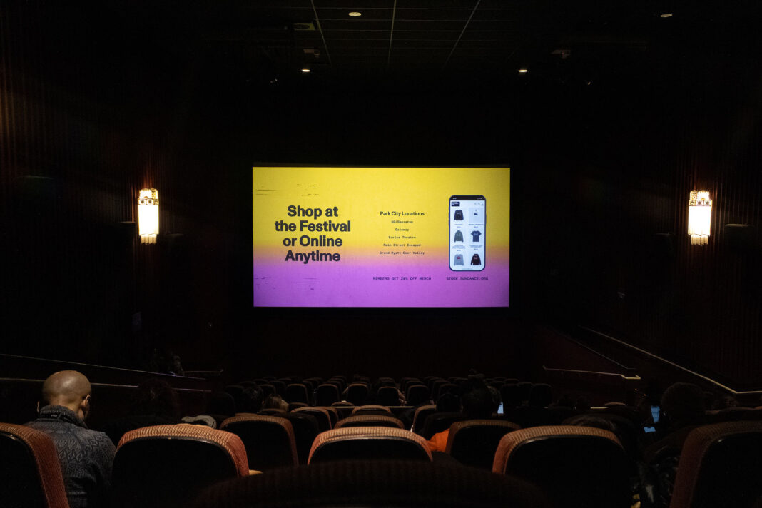 Landscape photo of a dark movie theatre with a Sundance Film Festival ad on screen and a few people sitting in seats