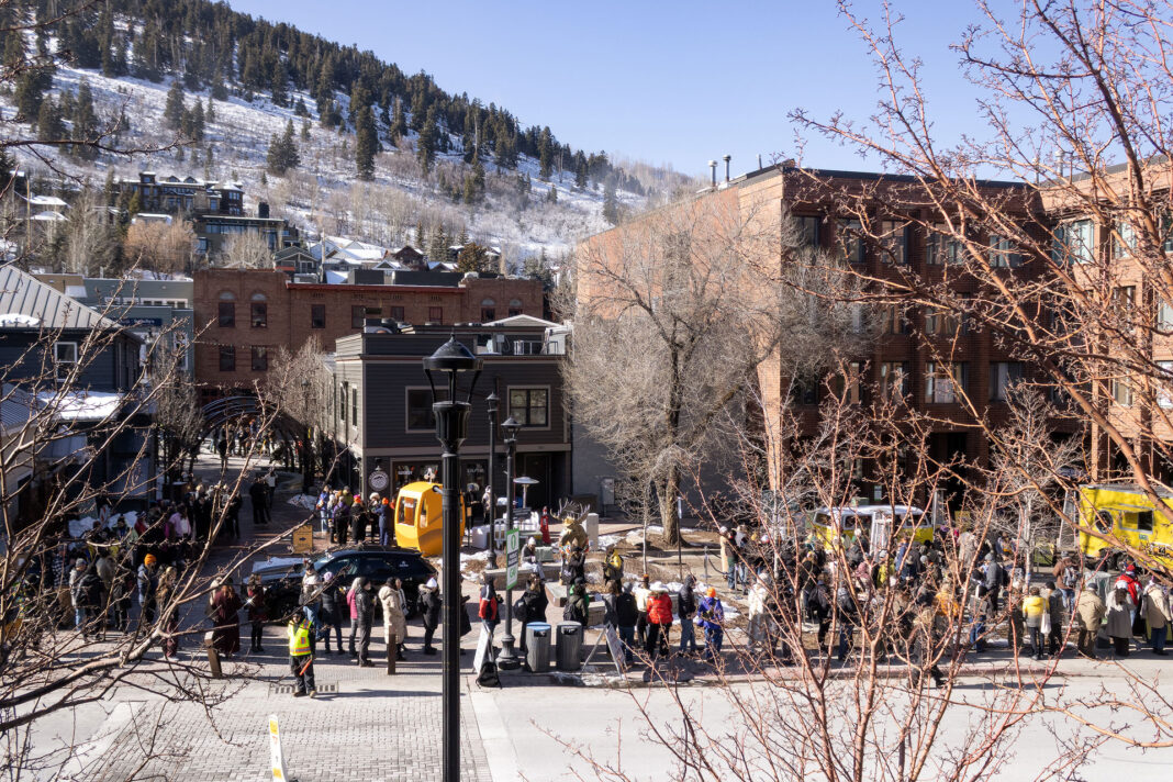 Landscape photo of a line of people, buildings, mountains, and a few dead branches making a frame. A single street light can be seen overlooking the event