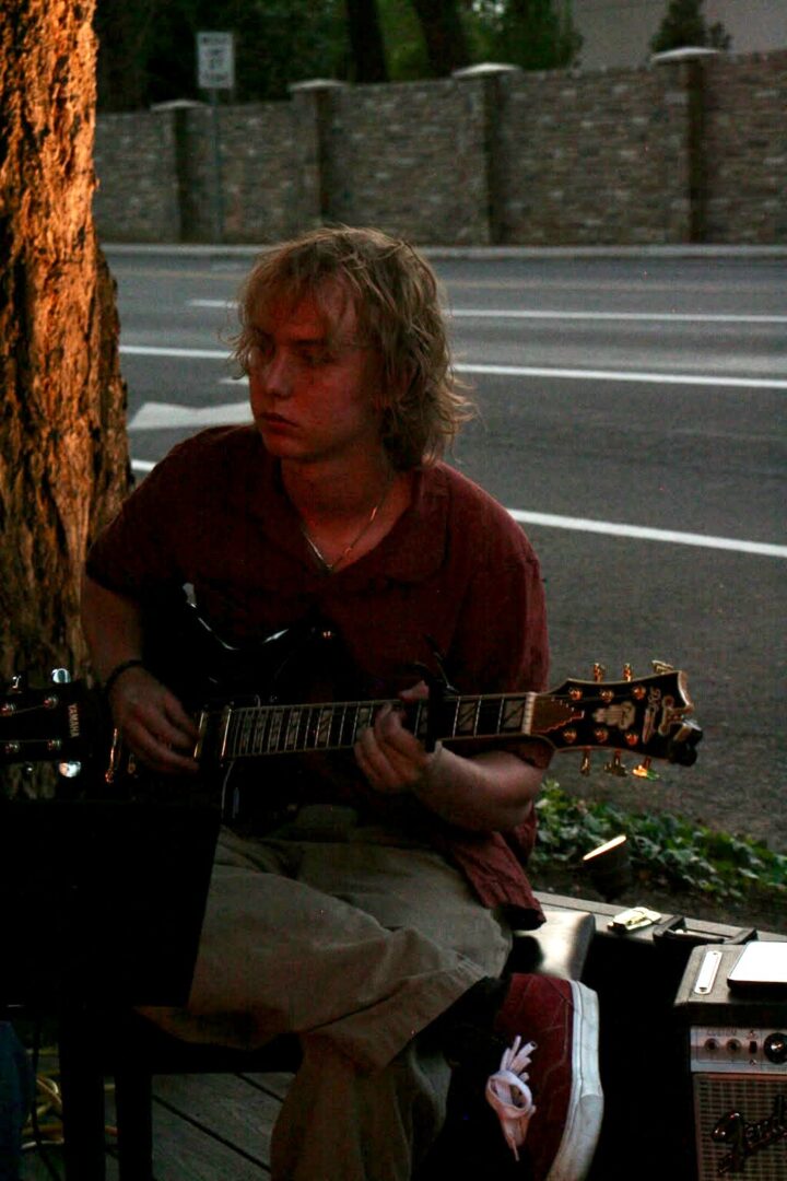 Trace playing a guitar on a sidewalk at dusk