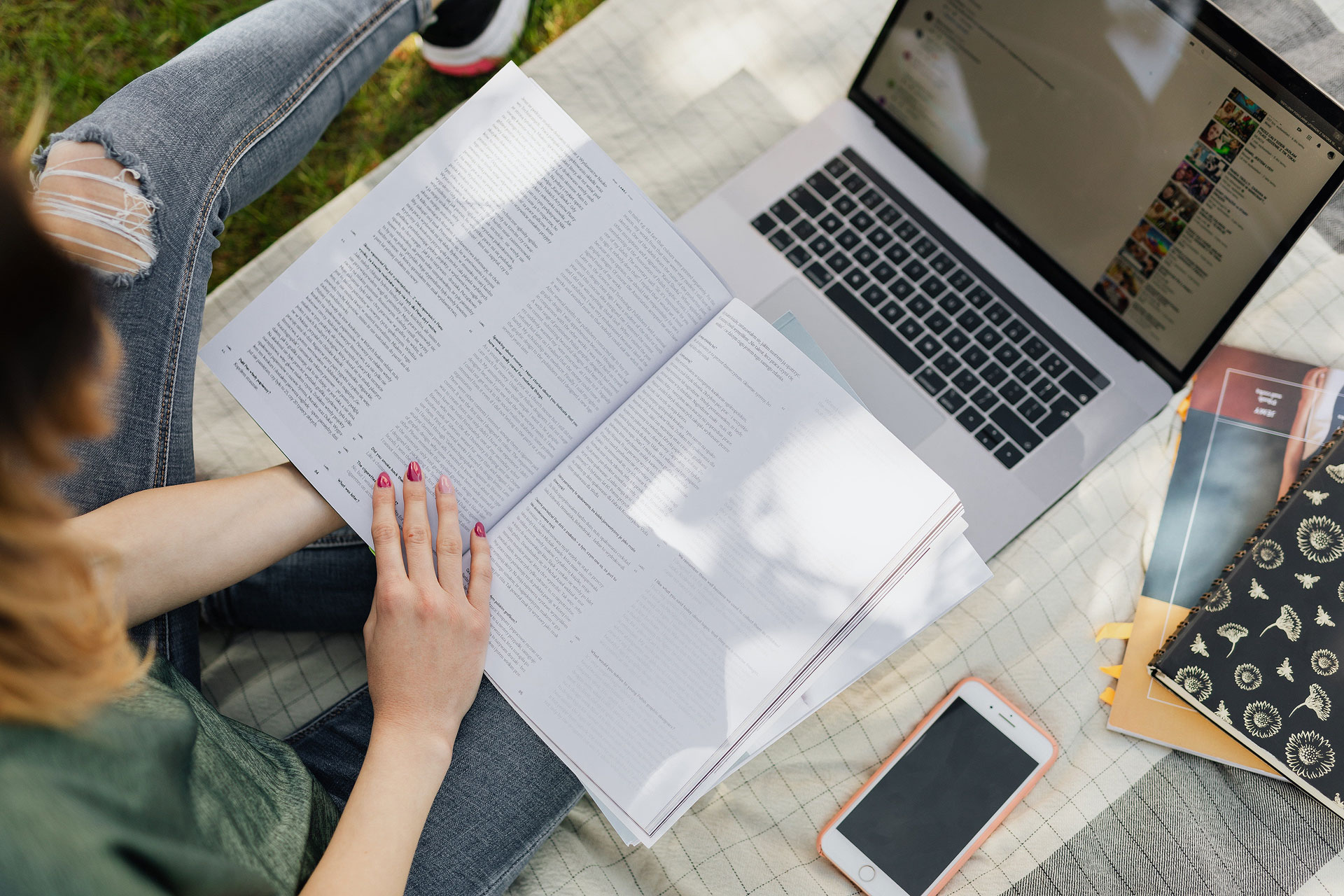 Woman reading an open book outside under shade with laptop and phone