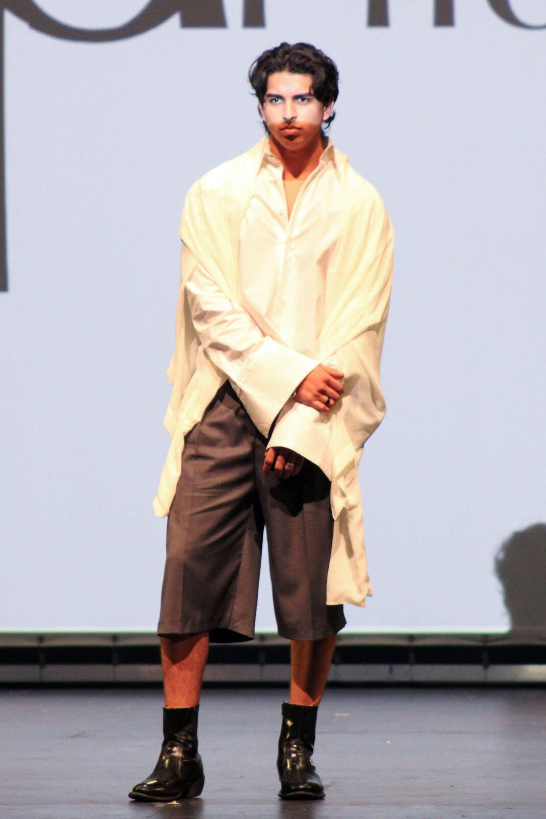 Model wearing an oversized white dress shirt, cutoff gray dress pants, and black boots on a stage