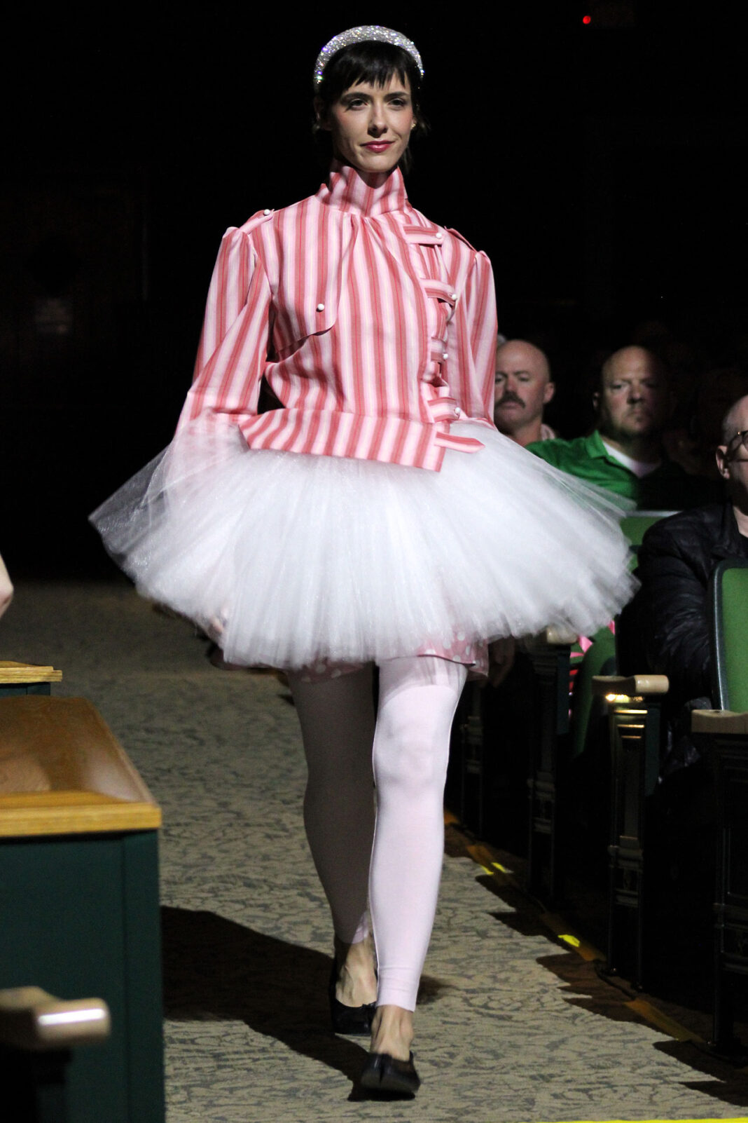 Model walking down an aisle wearing a sparkling head ornament, a pink and white striped top, and a white skirt and leggings