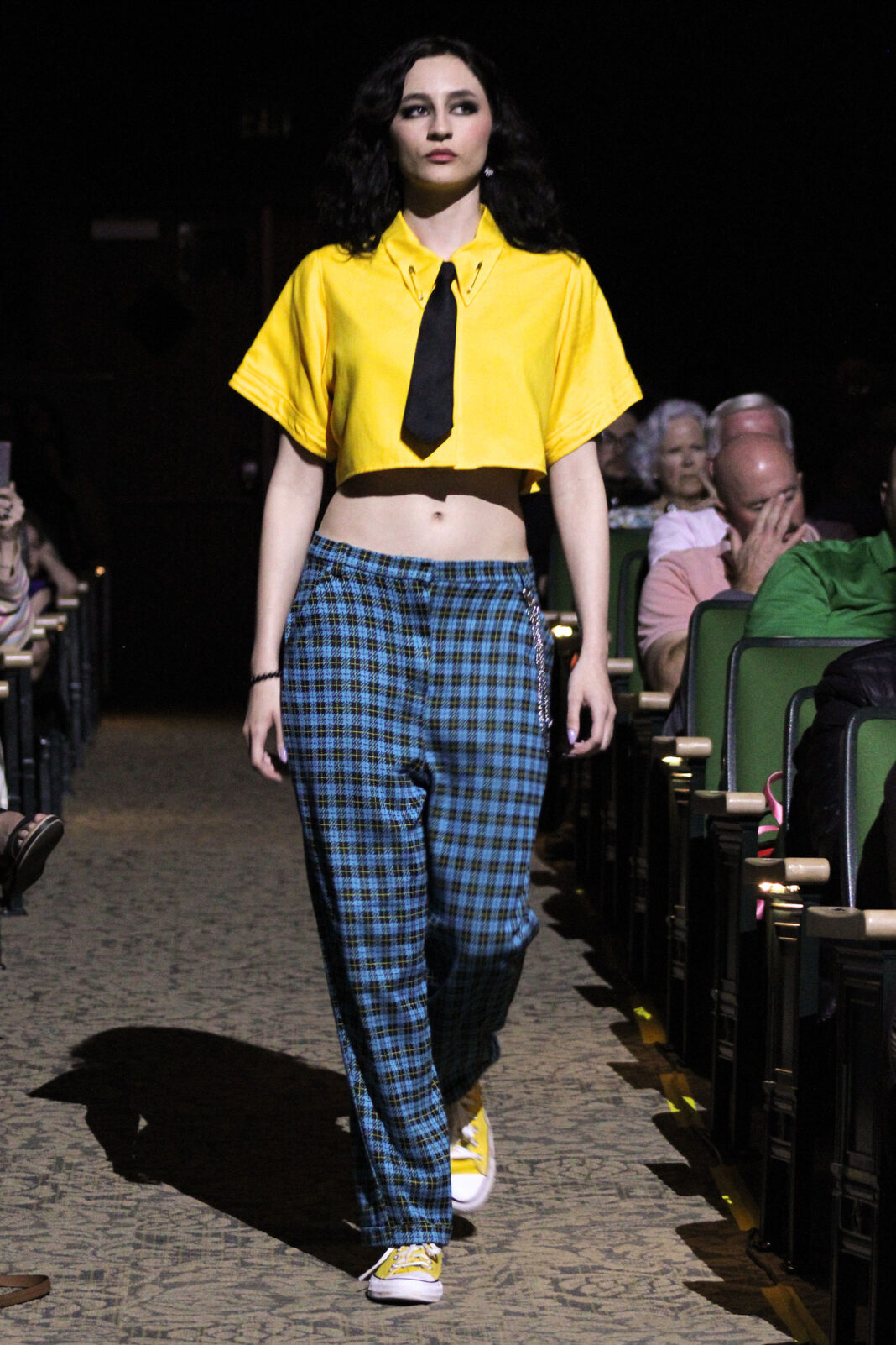 Model walking down an aisle wearing a yellow dress shirt crop top, a black tie, blue and black plaid pants, and yellow sneakers