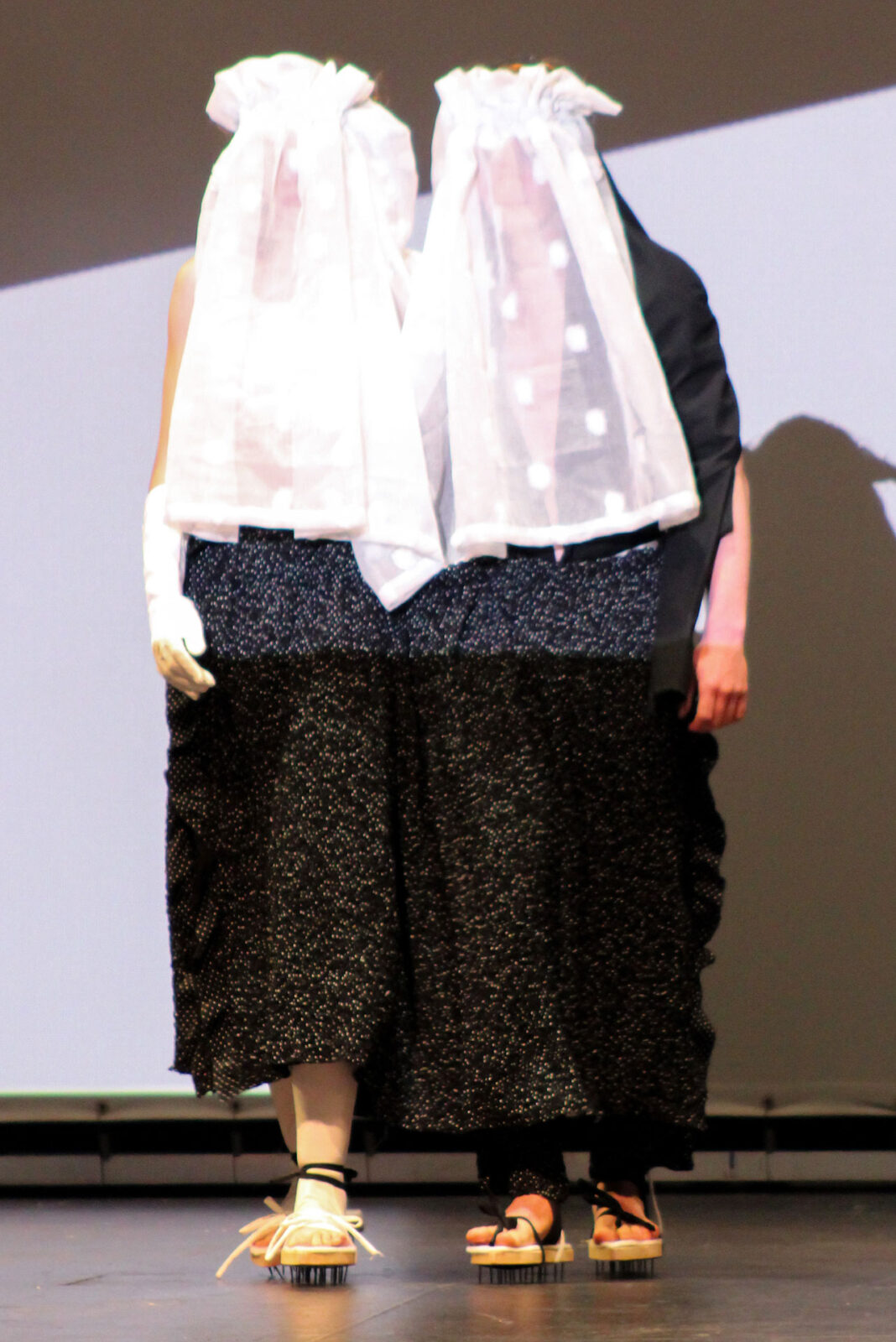 Two models on sage walking side by side wearing sandals with spikes on the bottom, a skirt, and matching headdresses