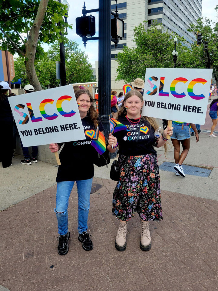 Mother and daughter holding Pride flags and SLCC signs while standing on a city corner
