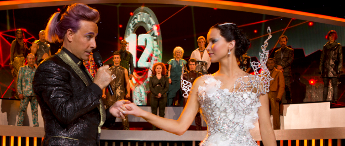 Movie review: No sparks flame ‘Catching Fire’ The film is once again caught between the realms of theatrics and realism, making for a moderate confusing time. Featured: Stanley Tucci (Left) reprising his role as Casear Flickerman, and Jennifer Lawrence (Right) reprising her role as Katniss Everdeen.