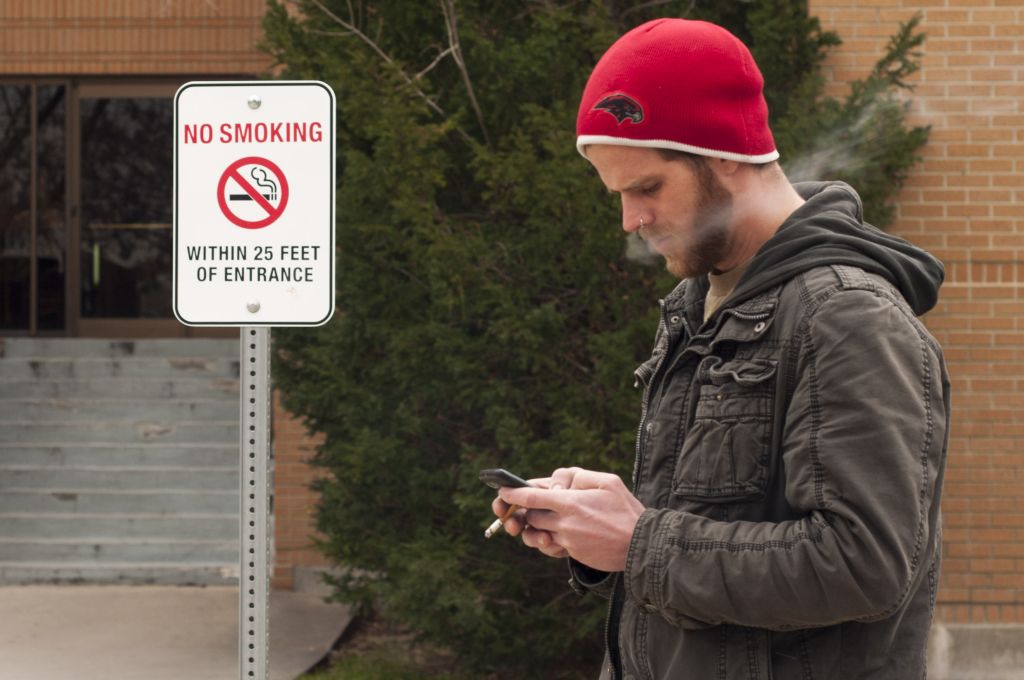 Health & Wellness proposes a campus wide ban on tobacco A student smoking a cigarette and texting in front of an entrance to the TB building. Right behind him is a sign that says no smoking within 25 feet of the entrance.
